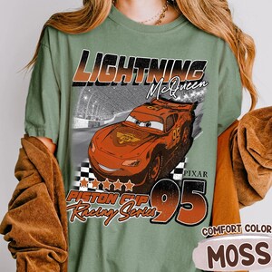 May include: Moss green t-shirt featuring Lightning McQueen from the Pixar Cars movie. The design includes the text "LIGHTNING McQueen", "PISTON CUP Racing Series", and the number "95". The shirt is labeled "COMFORT COLOR MOSS".