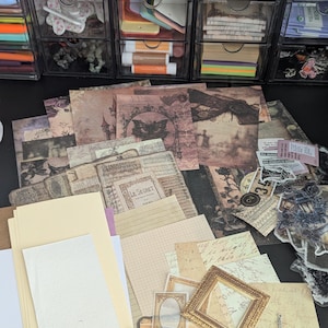Può includere: Una collezione di carta per scrapbook, elementi effimeri e abbellimenti in stile vintage. Include carte decorate con disegni antichi, cornici e piccoli ciondoli in metallo. La tavolozza dei colori presenta toni seppia, crema e tocchi di viola.