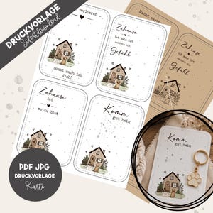 May include: A digital print template featuring four rectangular cards with rounded corners, each displaying a small house illustration, a dog, and German text. One card is shown with a gold-toned metal keychain featuring a paw print charm.