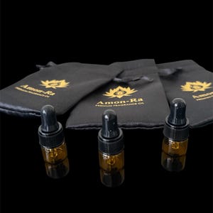 May include: Three small amber glass bottles with black dropper tops and gold labels that say "Amon-Ra Premium Fragrance Oil" are sitting on a black surface. The bottles are in front of three black velvet drawstring bags with the same logo.