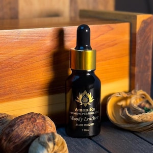 Op de afbeelding: Een zwarte glazen fles met een gouden dop en druppelaar. De fles is gelabeld met "Amon-Ra Premium Perfume Oil Woody Leather Made in Egypt 15ml".