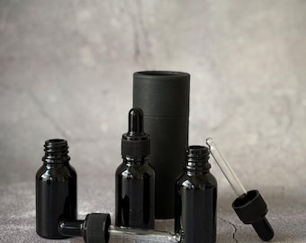 Może przedstawiać: Zestaw czarnych szklanych butelek z zakraplaczami i czarnymi zakrętkami oraz cylindryczny pojemnik. Jedna butelka ma przezroczysty szklany zakraplacz. Butelki są na szarej powierzchni, prawdopodobnie do kosmetyków.