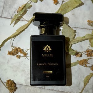 Puede incluir: Un frasco de perfume negro con una etiqueta rectangular con texto dorado que dice "Linden Blossom". La botella está etiquetada como "Amon-Ra Premium Perfume Oils" y "Made in Egypt". Hojas y flores secas rodean la botella.