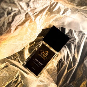 Puede incluir: Un frasco de perfume rectangular con tapa negra y etiqueta negra que dice "Vodka Smash" y "Scents Empire". El frasco es de 30 ml y está hecho a mano en EE. UU. El frasco está sobre una superficie dorada y blanca arrugada.