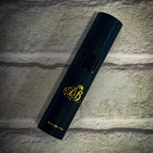 May include: A dark teal cylindrical perfume bottle with gold lettering and a gold crown design. The bottle has a black band and a black cap. The text on the bottle reads "AB" and "10 ml (0.3 fl oz)."