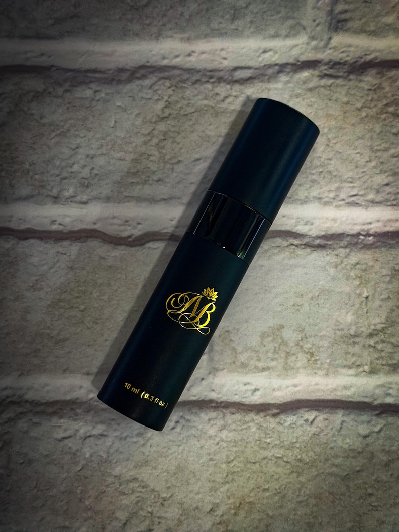 May include: A dark teal cylindrical perfume bottle with gold lettering and a gold crown design. The bottle has a black band and a black cap. The text on the bottle reads "AB" and "10 ml (0.3 fl oz)."