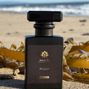 Puede incluir: Un frasco de aceite de perfume de pachulí negro de vidrio de Amon-Ra. El frasco está sentado en una playa de arena con algas en el fondo. La etiqueta es negra con letras doradas y presenta una flor de loto dorada.