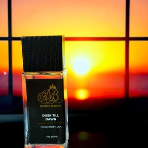 May include: A rectangular glass perfume bottle with a dark wooden cap. The black label reads "SCENTS EMPIRE" and "DUSK TILL DAWN", with "Handcrafted in USA" and "1.7oz (50ml)". The background is a vibrant sunset.