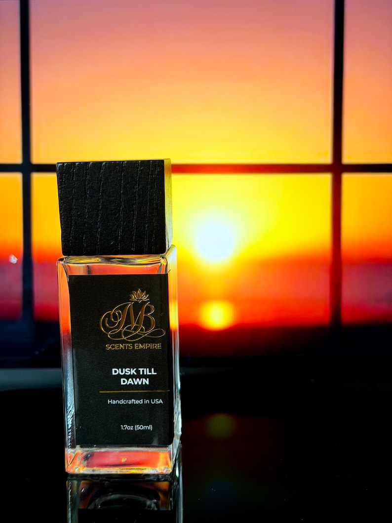May include: A rectangular glass perfume bottle with a dark wooden cap. The black label reads "SCENTS EMPIRE" and "DUSK TILL DAWN", with "Handcrafted in USA" and "1.7oz (50ml)". The background is a vibrant sunset.