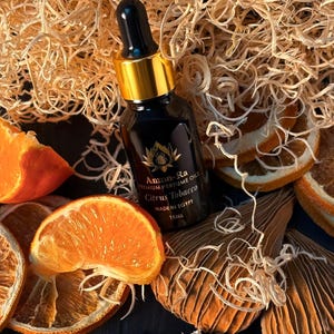 Op de afbeelding: Een klein donker glazen flesje met een gouden dop en druppelaar. Het flesje bevat een citrus-tabak geparfumeerde olie. Het etiket vermeldt "Amon Ra Premium Perfume Oils Citrus Tobacco Made in Egypt 15 ml". Het flesje is omringd door gedroogde sinaasappelschijfjes en houtsnippers.