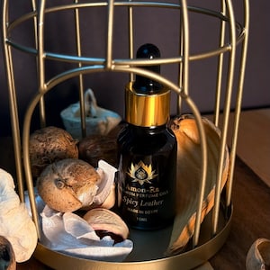 May include: A small, dark glass bottle with a gold cap and dropper. The bottle is labeled "Amon-Ra Premium Perfume Oils Spicy Leather Made in Egypt 15ML". The bottle is sitting inside a gold birdcage on a wooden surface.