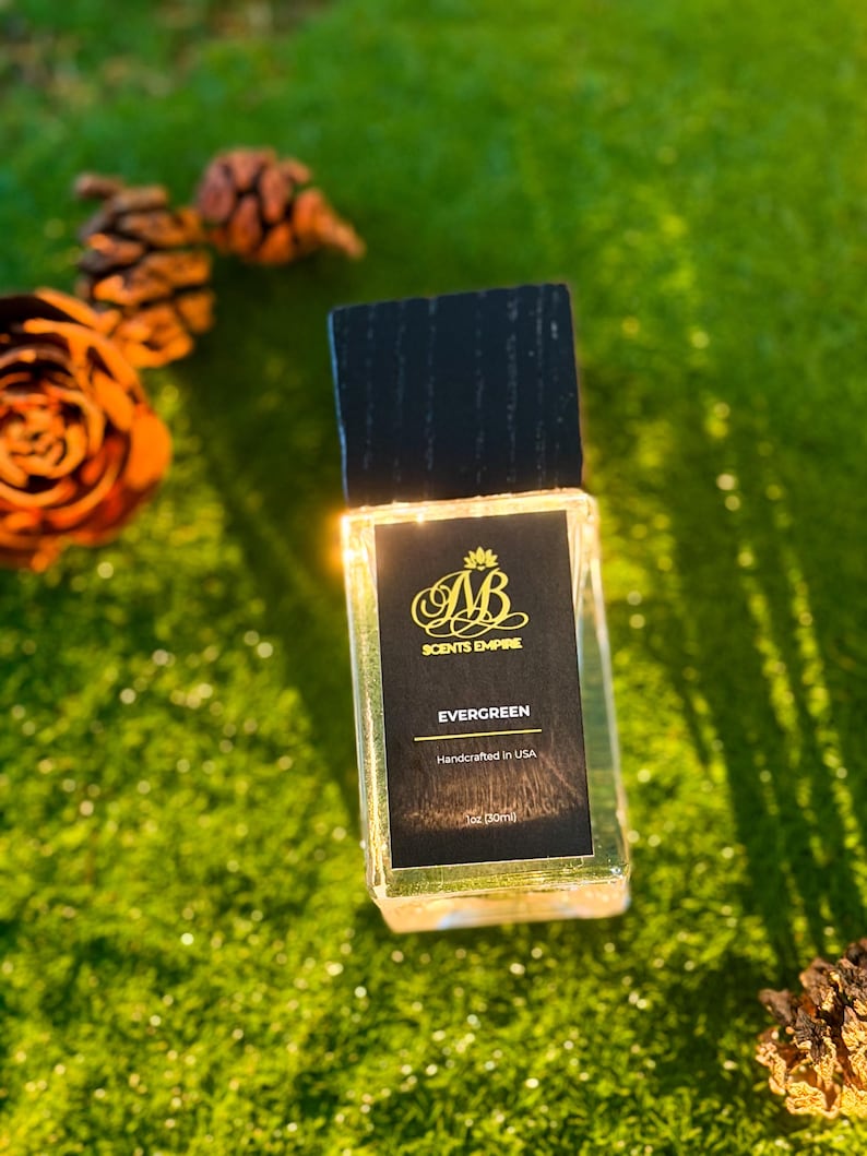 K&ouml;nnte beinhalten: Ein rechteckiger Glasparf&uuml;mflakon mit einem schwarzen Holzdeckel. Das schwarze Etikett zeigt das goldene Logo AB SCENTS EMPIRE, den Text EVERGREEN, Handcrafted in USA und 1oz (30ml). Der Flakon ist durchsichtig und zeigt die Fl&uuml;ssigkeit.