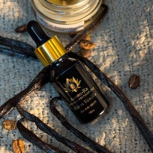 Puede incluir: Frasco de 15 ml de aceites de perfume premium Aom-Ra con aroma a vainilla cálida, con tapa dorada y etiqueta negra. El frasco está sobre una superficie gris con vainas de vainilla y granos de café.