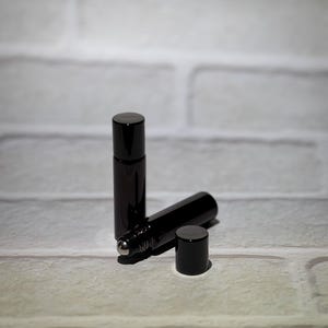 May include: Two black glass roll-on bottles with black caps. One bottle is upright, the other is lying on its side with the cap removed. The bottles have a silver rollerball applicator. The background is a white brick wall.