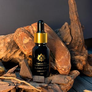 Puede incluir: Una botella de vidrio negro de aceite de perfume Amon-Ra Kalemat Oud con tapa y cuentagotas dorados. La botella es de 15 ml y está hecha en Egipto. La botella se exhibe sobre un lecho de virutas de madera y madera flotante.