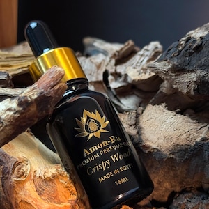 Może przedstawiać: Czarna szklana butelka ze złotą zakrętką i kroplomierzem. Butelka jest oznaczona etykietą "Amon-Ra Premium Perfume Oils Crispy Wood Made in Egypt 15ML".