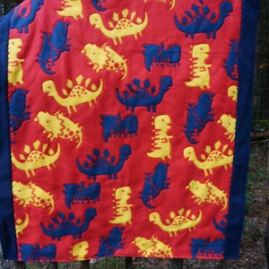 May include: Red blanket with a pattern of yellow and blue dinosaurs. The blanket has a dark blue border. The dinosaurs are various shapes and sizes, creating a playful design. This blanket is suitable for children.