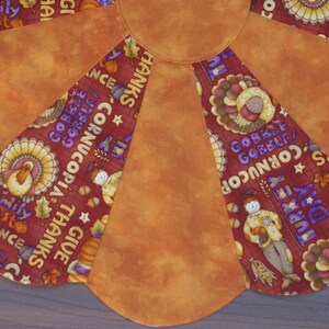 May include: A decorative, fan-shaped Thanksgiving table runner. The runner features a central orange section with alternating segments of patterned fabric. The patterned fabric includes turkeys, pumpkins, and text such as "Family Feast" and "Thanks".