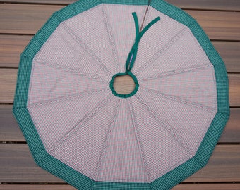 May include: A round Christmas tree skirt with a red and white checkered pattern. The skirt has a green border and a green tie. The skirt is made of fabric and is designed to go around the base of a Christmas tree.
