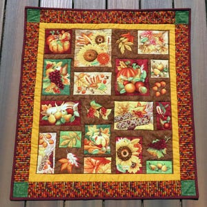 May include: A square autumn-themed quilt with a brown background and a yellow border. The quilt features various squares with images of pumpkins, sunflowers, leaves, grapes, and corn. The border is a mix of orange, brown, and yellow.