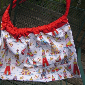 May include: A white tote bag with a red polka dot trim and handle. The bag features a colorful pattern of lighthouses, sailboats, and sea elements. The bag is made of fabric and is designed for carrying items.