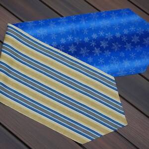 May include: Two decorative table runners. One features a blue background with white snowflakes. The other has a striped pattern in gold, blue, and gray. The runners are placed on a wooden surface.