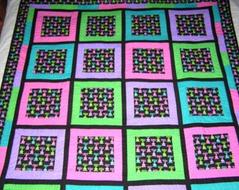 May include: A colorful quilt with a repeating pattern of squares. Each square is framed in black and contains a smaller square with a repeating pattern of black, pink, green, and blue figures. The quilt's border has a similar design.