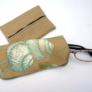 May include: Two eyeglass cases and a pair of glasses. One case is tan with a ribbed texture, the other is tan with a seashell pattern in blue and white. The glasses have a brown and white patterned frame.