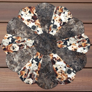May include: A circular, flower-shaped placemat featuring a central dark brown circle surrounded by eight petal-shaped sections. Each petal displays a collage of various dog breeds in shades of brown, black, and white. The placemat rests on a wooden surface.