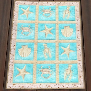 May include: A quilted wall hanging featuring a marine theme. The design includes alternating squares of light blue and beige, each adorned with starfish, crabs, lobsters, and seashells. The border is a patterned fabric with small, repeating motifs.