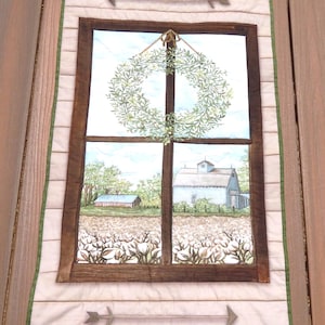 May include: A quilted wall hanging featuring a window scene with a wreath, a cotton field, and a farmhouse. The window is framed in brown, with arrows at the top and bottom. The border is white with a green trim.