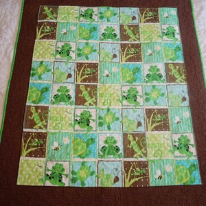 May include: A handmade quilt featuring a repeating pattern of green frogs, turtles, and lizards on a patchwork design. The quilt has a brown border with a green trim. The word "Green" is written in a cursive font on the quilt.