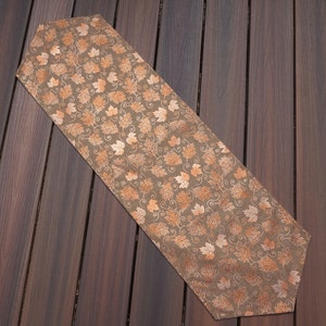 May include: A brown table runner with a repeating pattern of autumn leaves in shades of orange and cream. The runner is rectangular with a pointed end, perfect for a dining table or buffet. The fabric appears to be a linen or similar material.