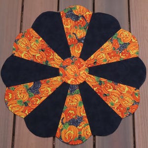 May include: A Halloween-themed placemat with a circular design. The placemat features alternating sections of black fabric and fabric printed with orange jack-o'-lanterns and black cats. The edges are scalloped, creating a decorative shape.