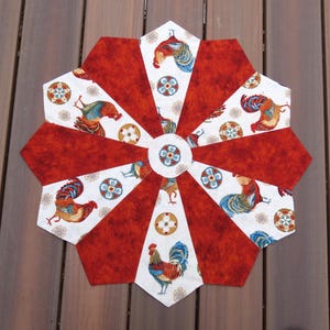 May include: A handmade, octagonal fabric trivet with a rooster theme. Alternating red and white panels radiate from a central floral design. The white panels feature rooster illustrations in shades of red, blue, and gold. The trivet is designed to protect surfaces from heat.