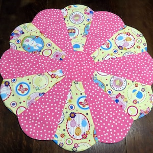May include: A flower-shaped quilted placemat with a pink and white polka dot pattern. The petals alternate with a light yellow fabric featuring Easter-themed illustrations of bunnies, eggs, and cupcakes. The center is a solid pink circle.