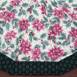 May include: A Christmas tree skirt with a poinsettia and holly pattern. The skirt features large pink poinsettias, green holly leaves, and red berries on a cream background. The border is dark green with a wreath pattern.
