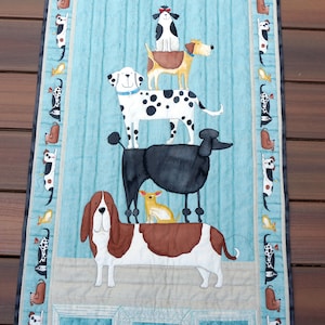 May include: A rectangular dog-themed quilt featuring a stack of various dog breeds, including a Basset Hound, Dalmatian, and Poodle. The quilt has a light blue background with framed dog portraits and a border with dog illustrations.