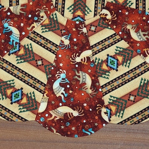 May include: Close-up of a fabric with a Native American design. The fabric features a repeating pattern of Kokopelli figures, geometric shapes, and stripes in shades of brown, tan, teal, and orange. The fabric is likely used for crafts or home decor.