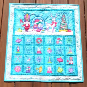 Puede incluir: Un calendario de adviento navideño acolchado con fondo azul claro y borde turquesa. Presenta imágenes festivas: cascanueces, bolas de nieve y árbol de Navidad. Cada día tiene un recuadro numerado.