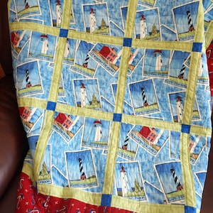 May include: A quilted blanket featuring a repeating pattern of lighthouse images on a blue background. The quilt has a green border and a red bottom border with an anchor design. The lighthouse images are labeled "Cape Hatteras" and "Fisgard Island".