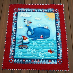 May include: A nautical-themed quilt featuring a blue whale wearing a sailor hat, a sailboat, and a seagull. The quilt has a red border with nautical flags and a sun in the corner. The background is a light blue with white clouds and ocean waves.