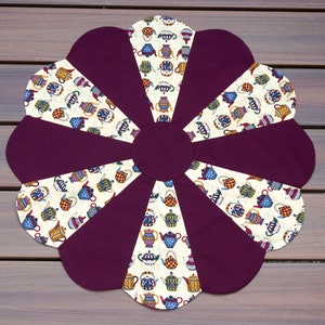 May include: A flower-shaped quilted placemat. The placemat features alternating sections of solid dark purple fabric and patterned fabric with colorful teapots on a cream background. The center is a solid dark purple circle.