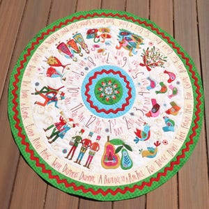 May include: A circular quilted fabric mat featuring the Twelve Days of Christmas theme. The design includes colorful illustrations of birds, dancers, and musicians arranged in a spiral with text labeling each day from 1st to 12th, finished with a green scalloped border.