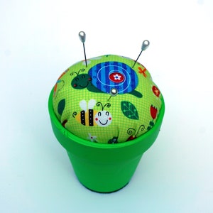 May include: A green painted flower pot holds a round pin cushion. The cushion is covered in a green fabric with a pattern of bees, turtles, and flowers. Two pins with silver heads are inserted into the cushion.