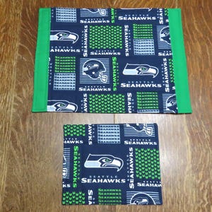 May include: Two Seattle Seahawks themed fabric placemats. The larger mat has a green border. Both mats feature a navy blue background with the team logo, helmet, and text "SEAHAWKS" in white and green.