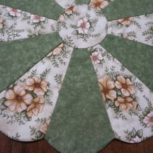 May include: A floral-patterned, quilted placemat in a fan shape. The design features alternating sections of solid green fabric and fabric printed with beige and brown flowers and green leaves. The center is a circle with the floral print.