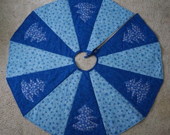 May include: A blue and light blue Christmas tree skirt with a snowflake and tree design. The skirt is pieced with alternating sections of solid and patterned fabric. The tree skirt has a center opening and tie closure.