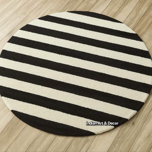 Op de afbeelding: Een ronde vloerkleed met afwisselende zwarte en cr&egrave;mekleurige strepen. Het vloerkleed is cirkelvormig en heeft een gedurfd, grafisch patroon. Het vloerkleed is gemaakt van een zacht materiaal en is geschikt voor een woon- of slaapkamer.