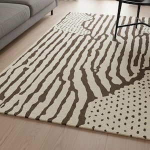 May include: A rectangular area rug with a cream-colored background and a brown abstract pattern. The design features vertical stripes, dotted sections, and irregular shapes. The rug is placed on a light-colored wooden floor.